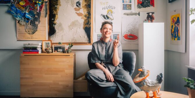 Eine junge Frau sitzt in einer Zimmerecke auf einem Sessel. An den Wänden im Hintergrund hängen verschiedene Wandbilder, im Vordergrund ist eine Taubenskulptur zu sehen. Foto: Christian Rückert