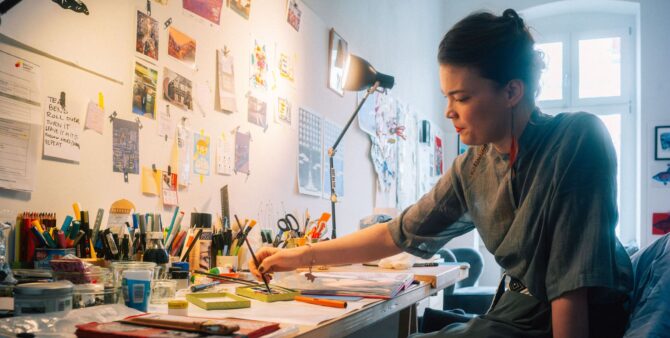 Eine junge Frau sitzt malend an einem Schreibtisch, auf dem zahlreiche Mal- und Bastelutensilien zu sehen sind. An der Wand hängen zahlreiche Bilder. Foto: Christian Rückert
