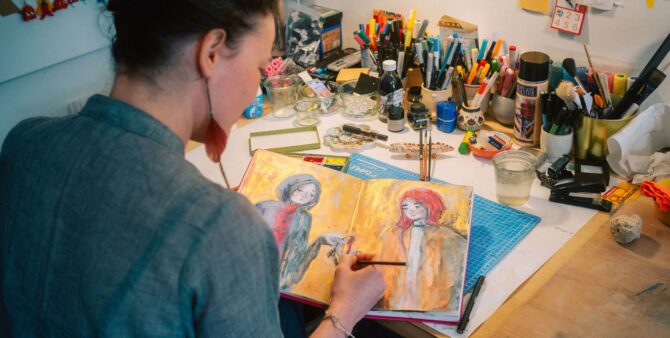Ansicht von hinten: Eine junge Frau sitzt an einer Arbeitsfläche mit zahlreichen Kunstmaterialien und malt in einem geöffneten Buch. Foto: Christian Rückert