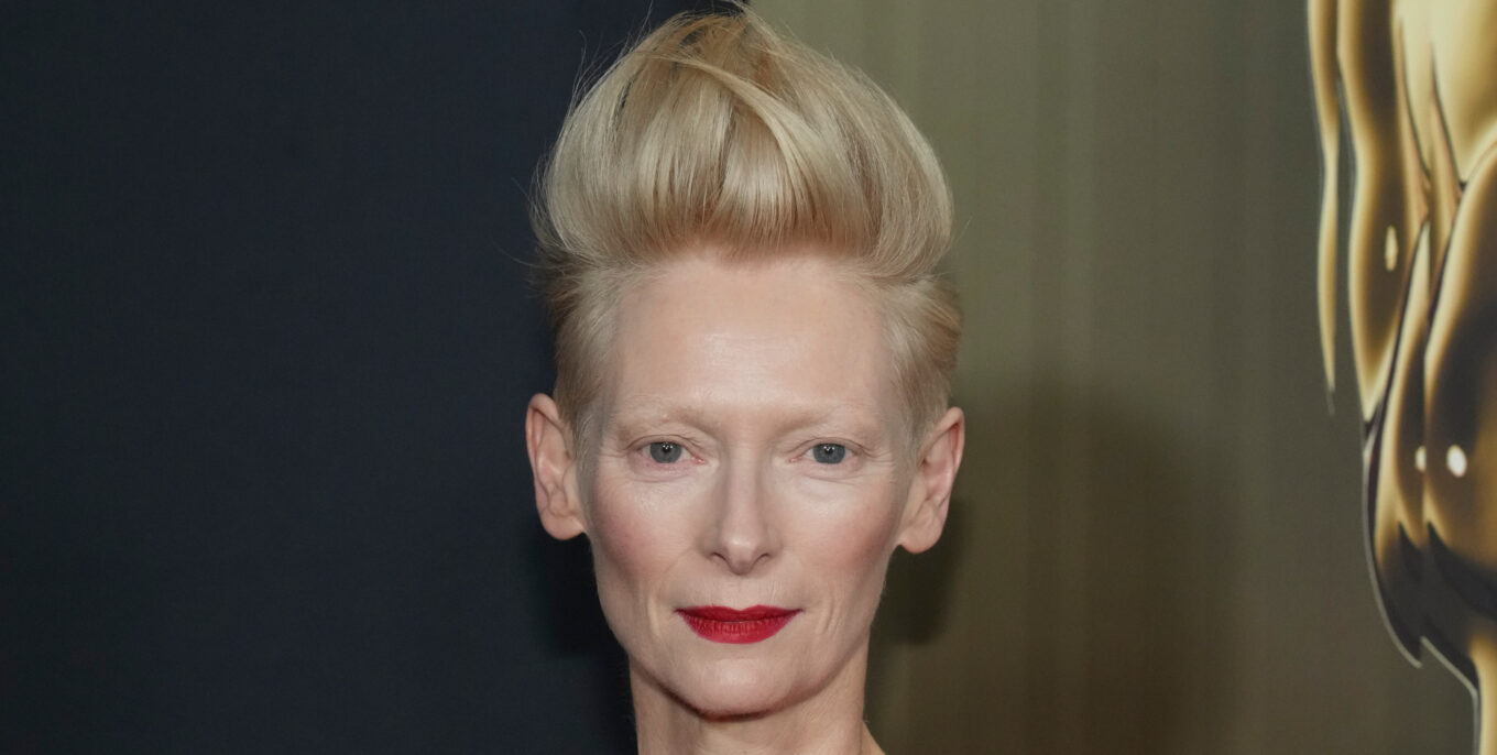 Nahaufnahme einer Frau mit blonden Haaren und rotem Lippenstift vor dunklem Hintergrund mit goldenem Detail.