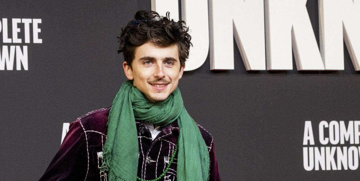 Mann mit grünem Schal und lila Jacke lächelt auf einem Event, im Hintergrund große weiße Schrift „A Complete Unknown“.