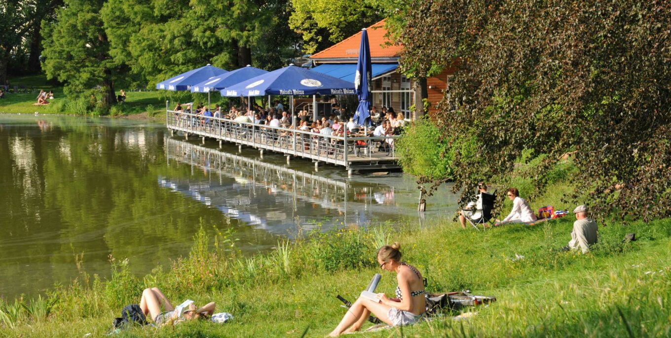 Seeidyll mit Biergarten und blauen Sonnenschirmen, Gäste sitzen auf Terrasse am Wasser