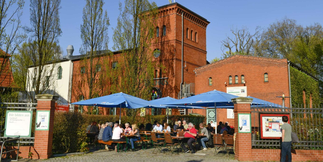 Biergarten vor rotem Backsteingebäude mit blauen Sonnenschirmen und Gästen bei Sonnenschein