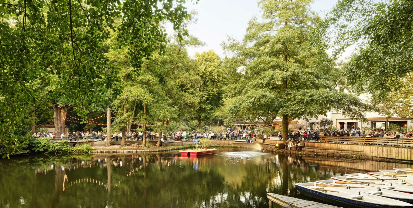 Großer Biergarten am See mit Booten, umgeben von Bäumen und Gästen unter Lichterketten