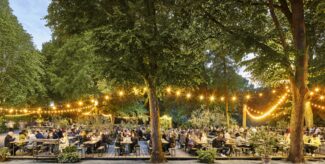 Romantischer Biergarten im Wald mit Lichterketten, vielen Gästen und Holzbänken am Abend