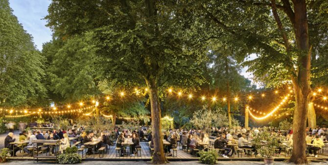 Romantischer Biergarten im Wald mit Lichterketten, vielen Gästen und Holzbänken am Abend