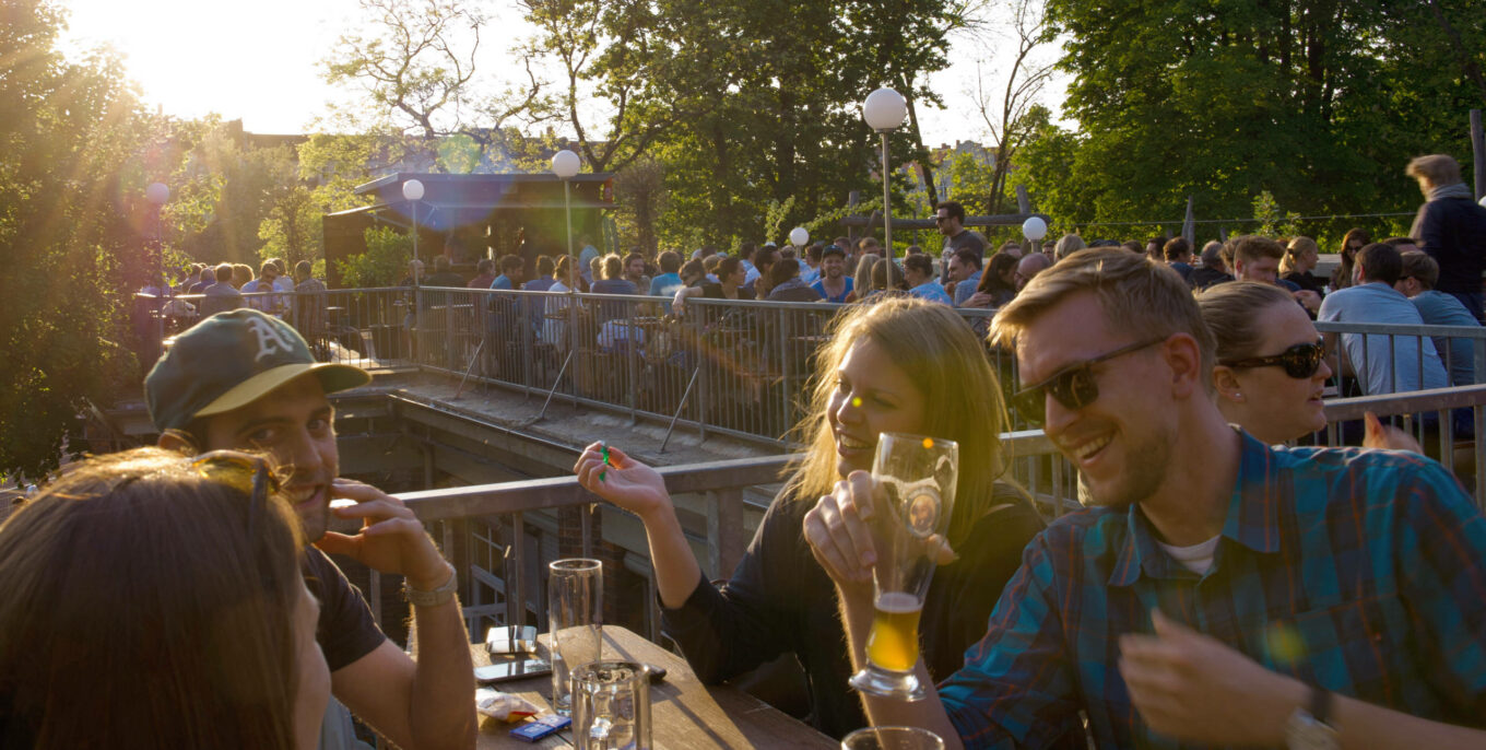 Freunde lachen und trinken Bier im sonnigen Biergarten, viele Gäste im Hintergrund