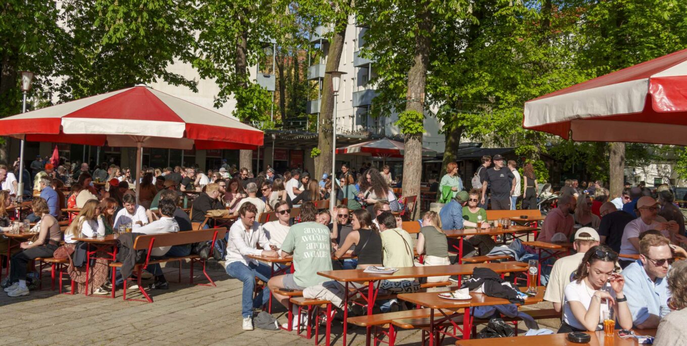 Lebhafter Biergarten mit rot-weißen Sonnenschirmen, langen Holzbänken und vielen Gästen