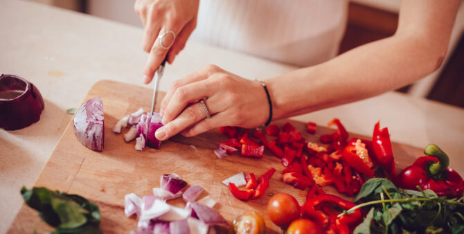 Hand schneidet rote Zwiebel auf Holzbrett, daneben Paprika, Tomaten und Basilikum.