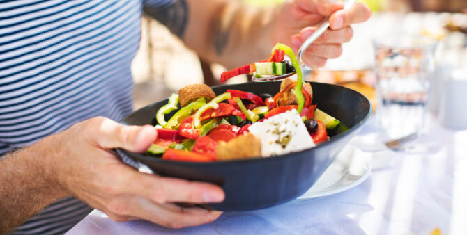 Mensch in gestreiftem Shirt isst bunten Salat mit Feta und Oliven im Sonnenlicht.