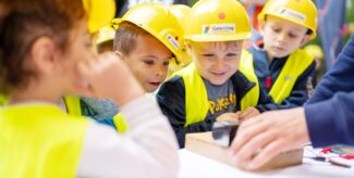 Kinder in gelben Bauhelmen und Warnwesten beobachten an einem Tisch, wie Erwachsenenhände etwas bearbeitet. Foto: Ralph Maak
