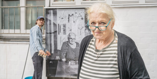 Eine ältere Frau mit Brille steht vor einem Haus. Im Hintergrund bringt ein Mann ein großes Schwarz-Weiß-Bild der Frau an einer Wand an. Foto: Felix Seyfert