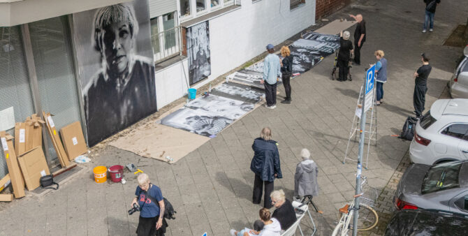 Eine Gruppe von Menschen auf einem Gehweg. Gemeinsam betrachten sie große Schwarz-Weiß-Fotografien, die auf nebeneinander auf dem Gehweg liegen und an eine Wand geklebt werden sollen. Foto: Felix Seyfert