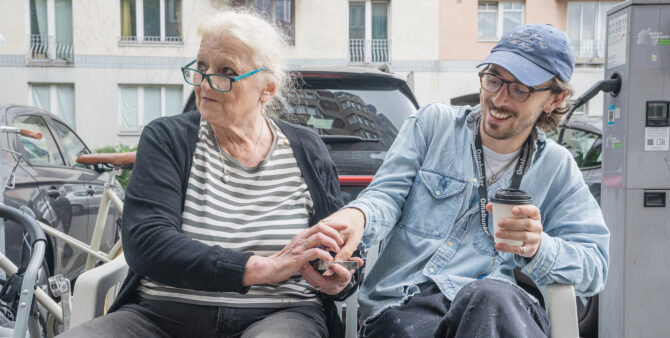 Eine ältere Dame und ein jüngerer Mann sitzen draußen auf einem Gehweg vor geparkten Autos. Foto: Felix Seyfert