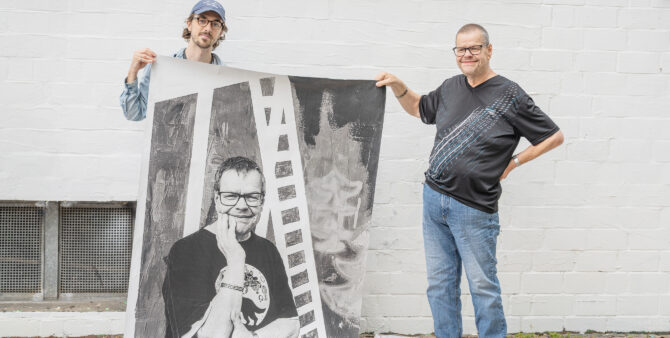 Zwei Männer halten gemeinsam eine große Schwarz-Weiß-Fotografie vor einer weißen Ziegelmauer. Foto: Felix Seyfert
