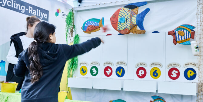 Ein junges Mädchen wirft einen Gegenstand auf eine Recycling-Wand mit bunten Fischen und verschiedenen Zeitangaben für den Abbau von Materialien. Foto: Felix Seyfert