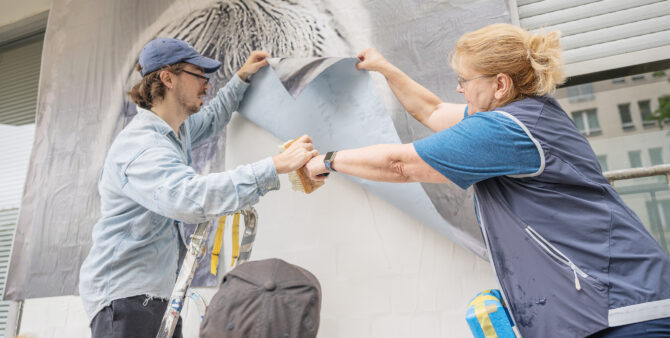 Ein Mann und eine Frau stehen auf Leitern und kleben gemeinsam ein großes Bild an eine Wand. Foto: Felix Seyfert