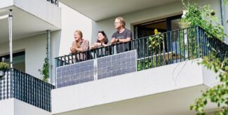 Zwei Frauen und ein Mann stehen auf einem Balkon in der Sonne und lehnen über der Brüstung. An der Außenseite des Geländers hängen zwei Photovoltaik-Paneele. Foto: Andreas Labes