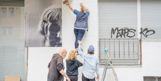 Vier Personen arbeiten zusammen, um ein großes Wandbild an eine Wand zu kleben. Foto: Felix Seyfert