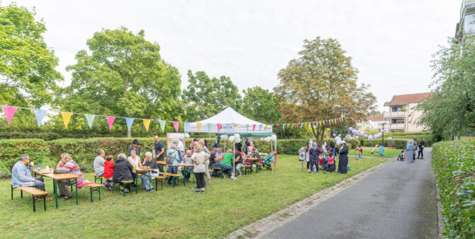Menschen sitzen und stehen auf einer grünen Wiese bei einem Fest unter einem weißen Pavillon mit bunter Wimpelkette. Foto: Felix Seyfert