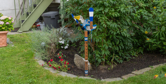 Eine hölzerne Ente auf einem Pfahl als dekorative Wetterfahne im Garten, umgeben von Blumen und Sträuchern. Foto: Felix Seyfert