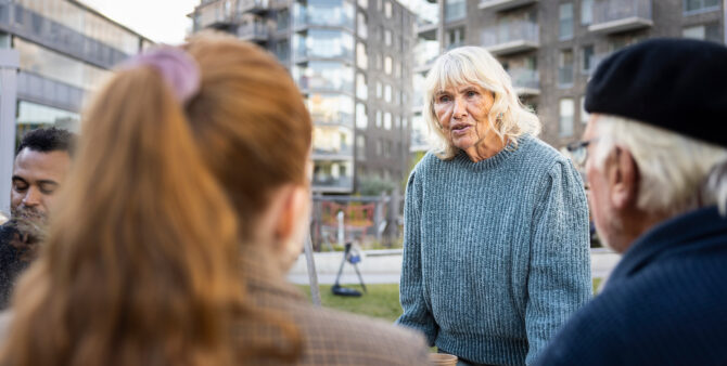 Ältere Frau spricht achtsam mit Nachbarn im Freien, umgeben von Wohnhäusern.