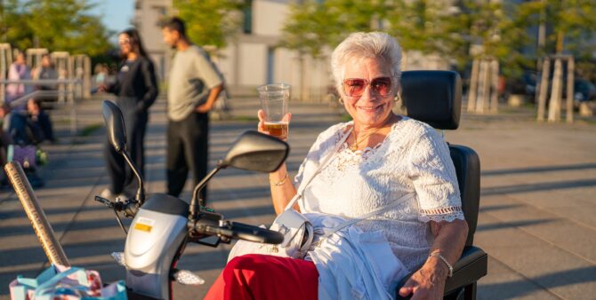 Aufnahme im Freien: Eine ältere Dame mit Sonnenbrille sitzt in einem elektrischen Seniorenmobil, hält ein Getränk in der Hand und lächelt in die Kamera. Foto: Ralph Maak