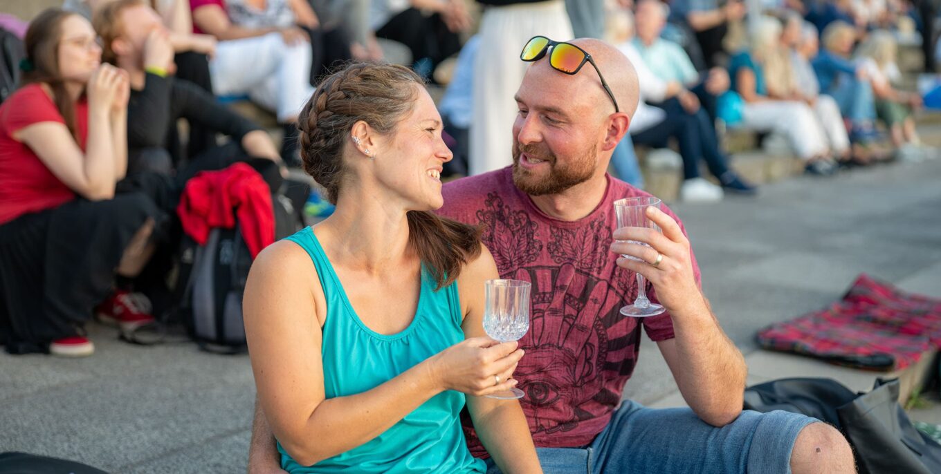 Ein junges Paar sitzt auf einer Treppenstufe im Freien und lächelt sich an. Beide halten ein Glas in der Hand. Foto: Ralph Maak