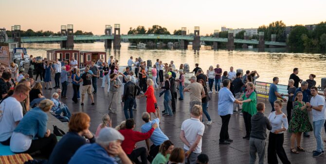 Zahlreiche Menschen bei einer Tanzveranstaltung auf einem Steg. Im Hintergrund ist ein großer Fluss samt einer Brücke zu sehen. Foto: Ralph Maak