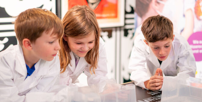 Drei Kinder in weißen Laborkitteln schauen konzentriert auf einen Tisch im Vordergrund. Foto: Ralph Maak