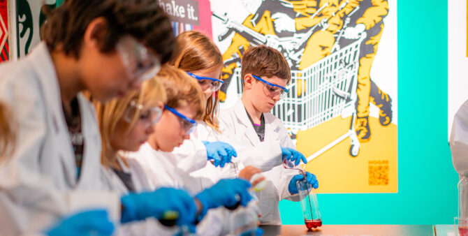 Mehrere Kinder in weißen Laborkitteln und blauen Handschuhen führen experimentelle Tätigkeiten aus, während sie ein Behältnis mit roter Flüssigkeit befüllen. Im Hintergrund ist ein farbiges Kunstwerk zu sehen. Foto: Ralph Maak