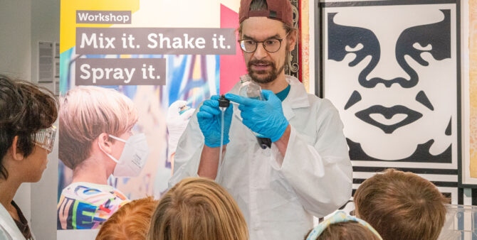 Ein Mann in einem weißen Laborkittel referiert bei einem Workshop vor Kindern. Foto: Ralph Maak