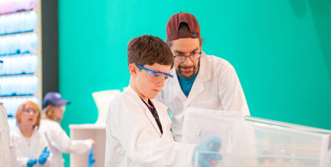 Eine Junge sprayt unter Aufsicht eines Erwachsenen mit einer Sprühdoese in einen großen durchsichtigen Behälter. Beide tragen weiße Laborkittel. Im Hintergrund sind weitere Kinder bei der Arbeit zu sehen. Foto: Ralph Maak