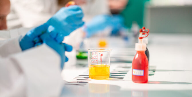 Detailaufnahme eines Gelben Bechers und einer roten Flasche auf einem Tisch mit Laborutensilien, umgeben von blauen Handschuhen. Foto: Ralph Maak
