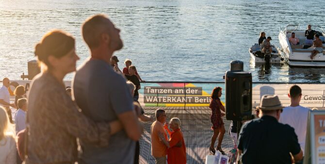 Mehrere Personen bei einer Tanzveranstaltung im Freien. Im Hintergrund ist ein Transparent an einer Uferpromenade zu sehen. Aufschrift: Tanzen an der WATERKANT Berlin. Foto: Ralph Maak