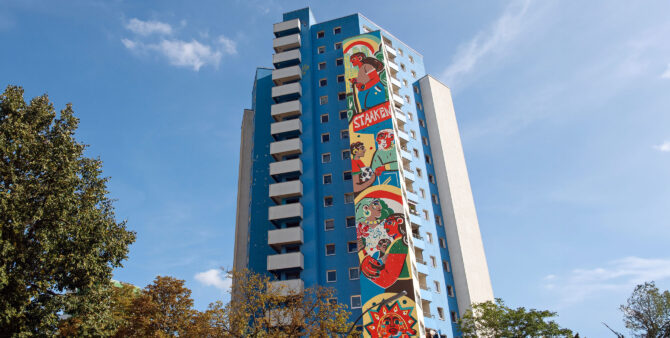 Drohnenaufnahme: Ein blau-weißes Hochhaus, an dem ein buntes, 50 Meter hohes Wandbild prangt. Foto: Jennifer Sanchez