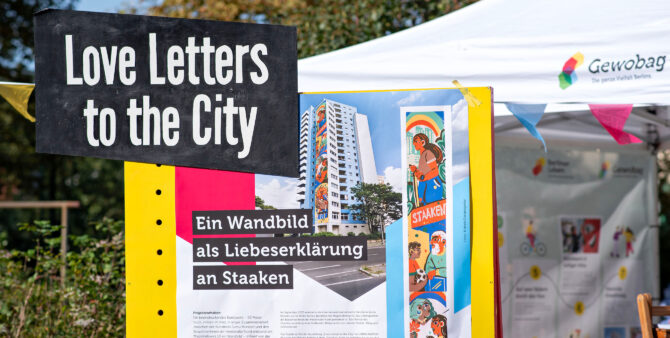 Detailaufnahme: An einem Gewobag-Stand auf einem Quartiersfest steht eine Erklärtafel mit der Aufschrift "Love Letters to the City - Ein Wandbild als Liebeserklärung an Staaken". Foto: Jennifer Sanchez