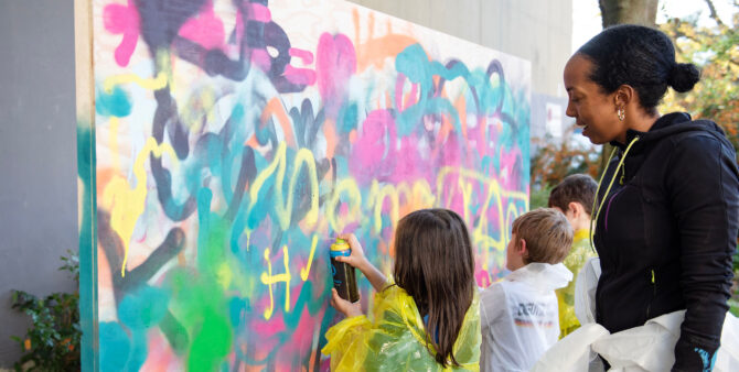 Drei Kinder besprühen eine Leinwand mit Graffiti, eine Frau steht dahinter und schaut zu. Foto: Jennifer Sanchez