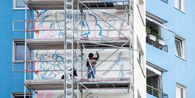 Ansicht von hinten: Eine Frau steht auf einem Baugerüst und bemalt eine Häuserfassade. Foto: Jennifer Sanchez