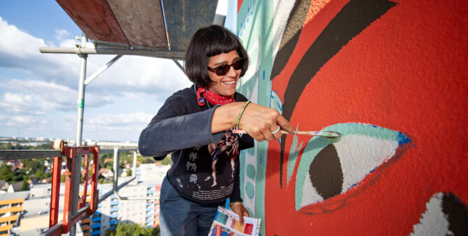 Eine lächelnde Frau mit Sonnenbrille mal mit einem Pinsel ein Kunstwerk auf eine Häuserwand. Dabei steht sie auf einem Baugerüst. Foto: Jennifer Sanchez