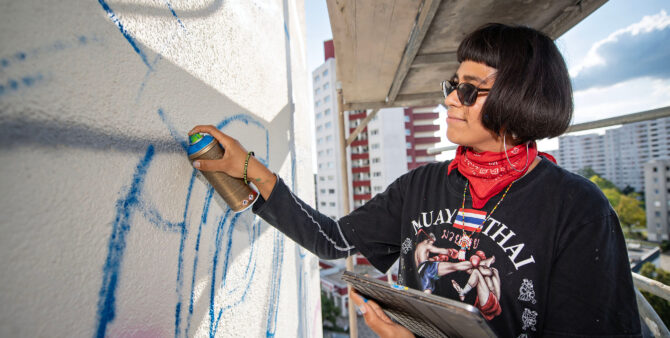 Eine Frau mit Sonnenbrille sprayt mit eine Sprühdose ein Graffito an eine Häuserwand. Dabei steht sie auf einem Baugerüst. Foto: Jennifer Sanchez