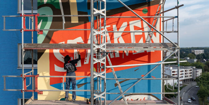 Großaufnahme: Eine Frau steht auf einem Baugerüst und bemalt eine Häuserfassade. Zu lesen ist das Wort "Staaken". Foto: Jennifer Sanchez