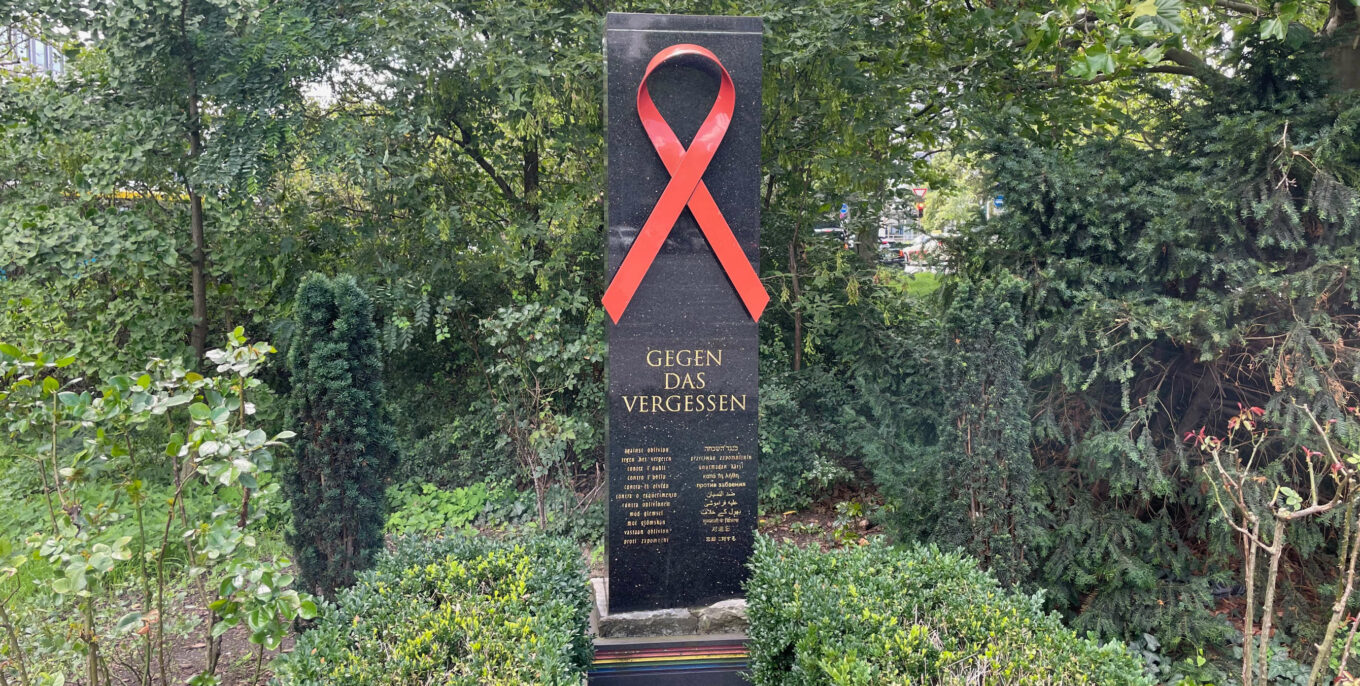 Gedenkstein mit roter AIDS-Schleife und Inschrift "Gegen das Vergessen".