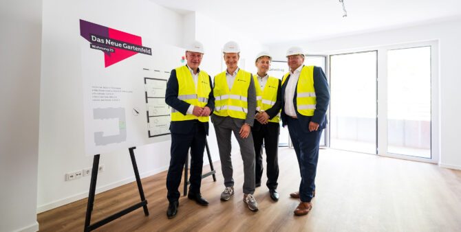 Vier Männer posieren in einer Musterwohnung neben einer großen Tafel mit dem dazugehörigen Grundriss. Foto: City-Press GmbH