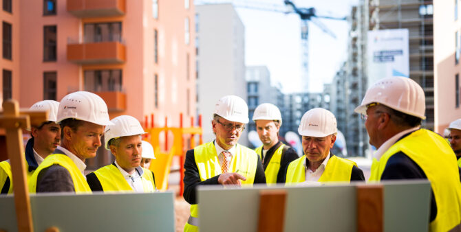 Eine Gruppe Männer mit Bauhelmen und gelben Leuchtwesten steht auf einer Baustelle und blickt auf Infotafeln. Einer der Männer erklärt. Foto: City-Press GmbH
