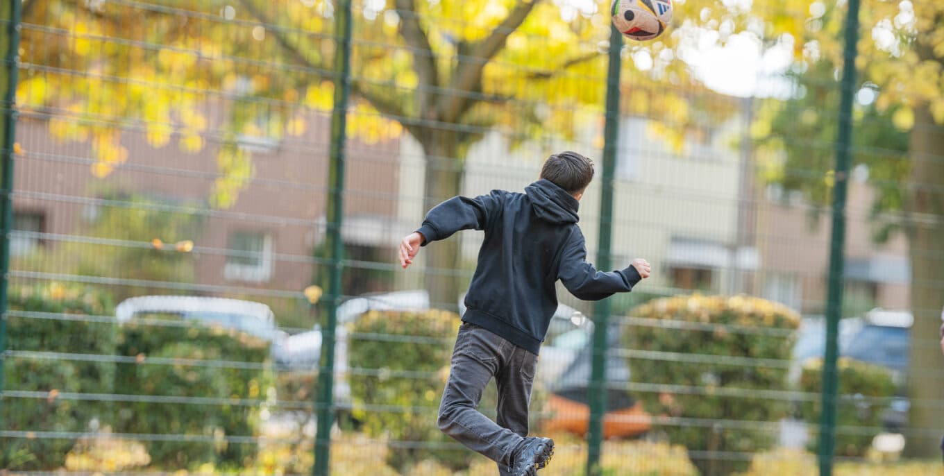 Ansicht von hinten: Ein Junge in schwarzem Kapuzenpullover bei einem Kopfball in einem Fußballkäfig. Foto: Felix Seyfert