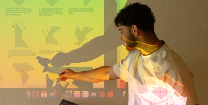 Momentaufnahme aus einem Workshop: Ein Schüler im Teenageralter steht vor einer Wand, auf die ein Beamer ein Bild wirft. In seiner Hand hält er ein gefaltetes Stück Papier, mit dem er ein Schattenmotiv kreiert. Foto: Ludger Paffrath