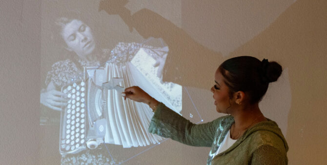 Momentaufnahme aus einem Workshop: Eine Schülerin im Teenageralter steht vor einer Wand, auf die ein Beamer ein Bild wirft. In ihrer Hand hält sie ein gefaltetes Stück Papier, mit dem sie ein Schattenmotiv kreiert. Foto: Ludger Paffrath