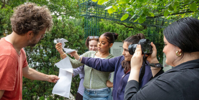 Momentaufnahme aus einem Workshop: Eine Gruppe von Schülerinnen im Teenageralter stehen im freien und halten gefaltete Papierstücke hoch. Eine weitere Person fotografiert die Aktivität. Foto: Ludger Paffrath