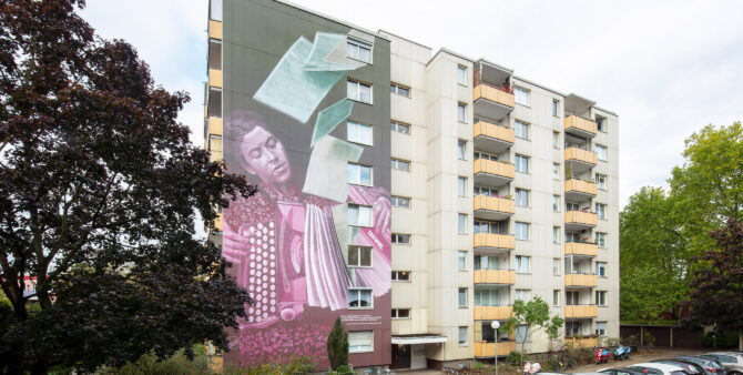 Ein Hochhaus mit einer bemalten Fassade. Darauf zu sehen: Eine akkordeonspielende Frau, deren Instrument sich bei genauerem Hinsehen als Schreibmaschine entpuppt. Foto: Ludger Paffrath